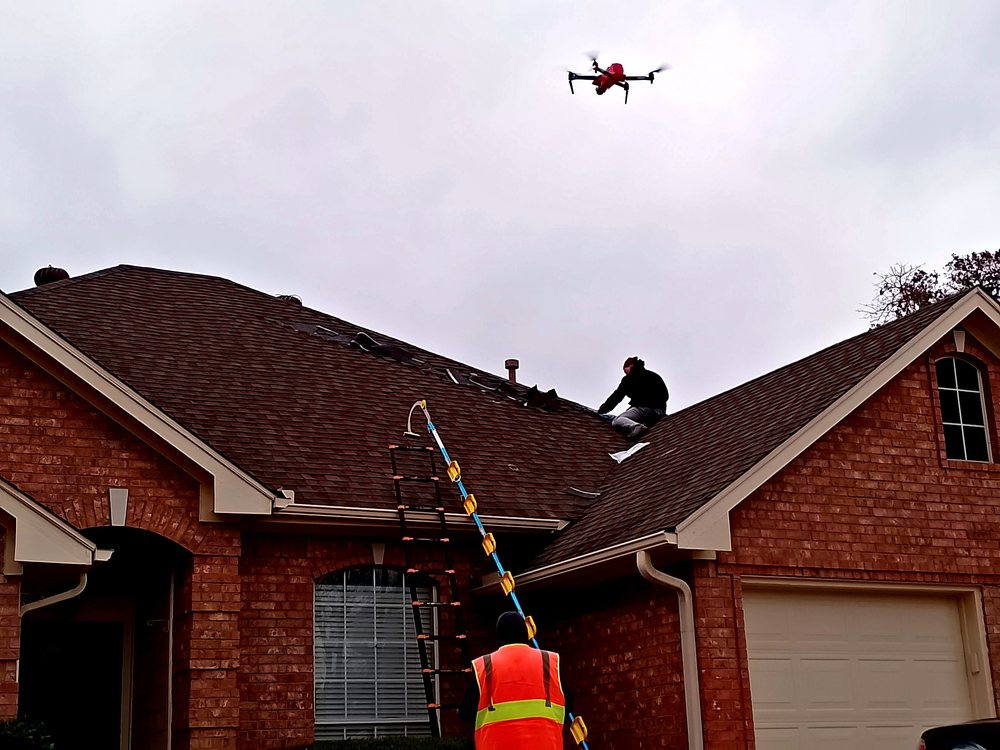 A drone hovering above a brick home's roof while an inspector climbs a ladder to the eave